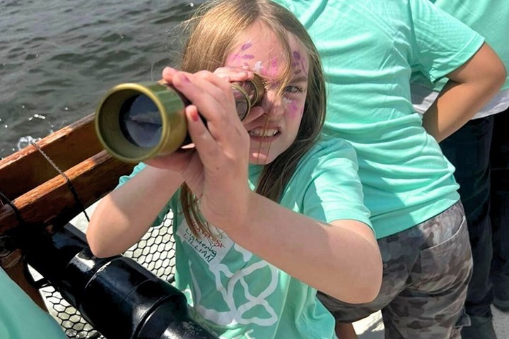 A child wearing a teal summer camp t-shirt and pink face paint looks through a telescope. Camp counselors stand behind them.