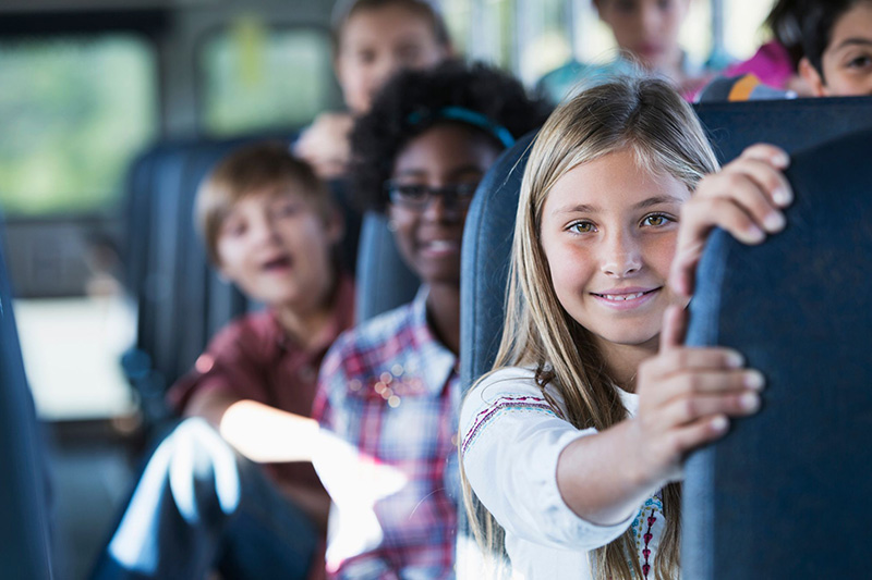 A young girl on a bus smiling with other children smiling behind her.