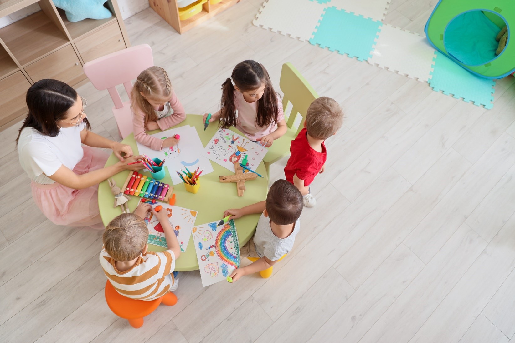 A bird’s eye view of a teacher kneeling at a table while 5 children color during child care. 