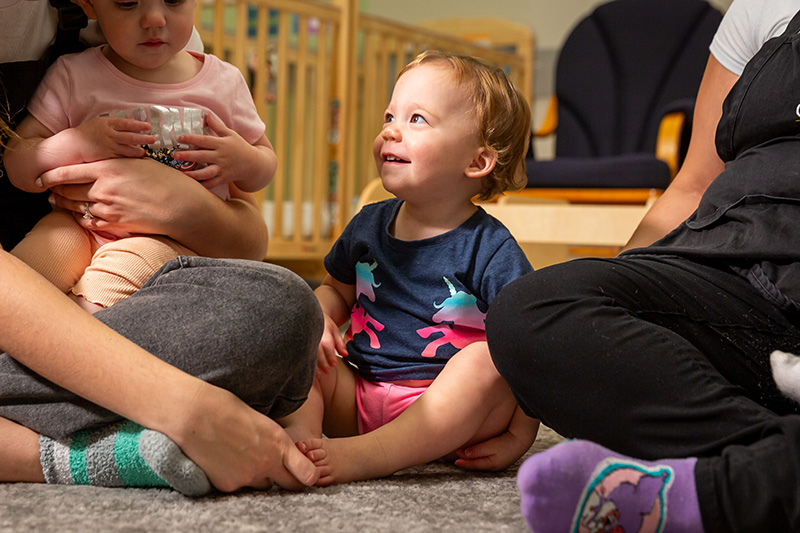 A baby sits on the floor and smiles at their caregiver, who is sitting beside them while holding another baby in their lap.