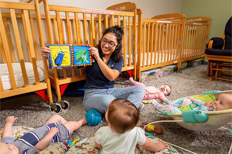 A caregiver sits in front of a row of cribs as they read a book aloud. Four infants sit and lie on the floor beside them.