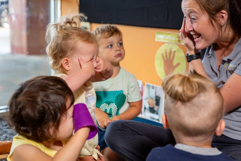 A Celebree child care giver sitting with toddlers during circle time and pointing to her eyes while a little girl covers her eyes with her fingers. 