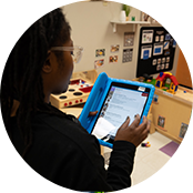 A child care worker uses an electronic tablet with a blue case to write progress reports for their preschool students.