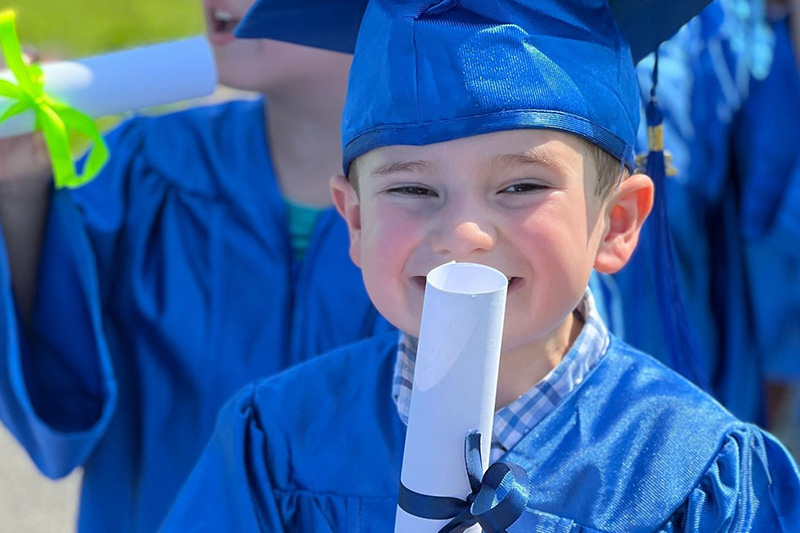 A preschooler smiles at the camera while holding their diploma and wearing a blue cap and gown on graduation day.