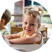 A toddler with blonde hair in pigtails sits at a white table and smiles at the camera.