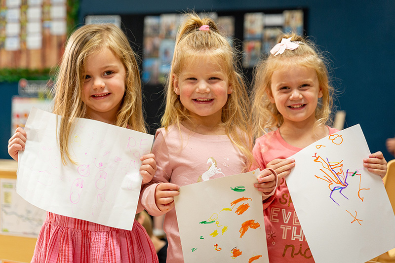 Three preschoolers smile at the camera as they show off the drawings they created during the school day.